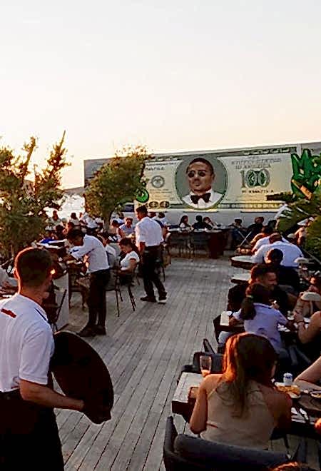 Nusr-Et Yalıkavak’ta Salt Bae duvar resmi ve gün batımı manzaralı deniz kenarında açık oturma alanı ile canlı bir yemek ortamı.